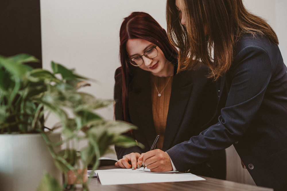 Office assistant sitting at a desk witnessing another person signing a document.